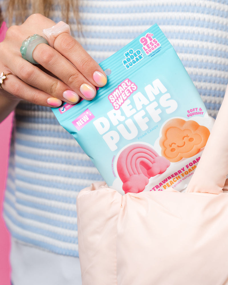 Hand pulling a blue bag of SmartSweets Dream Puffs out of a pink tote bag, showing strawberry & peach candy images.