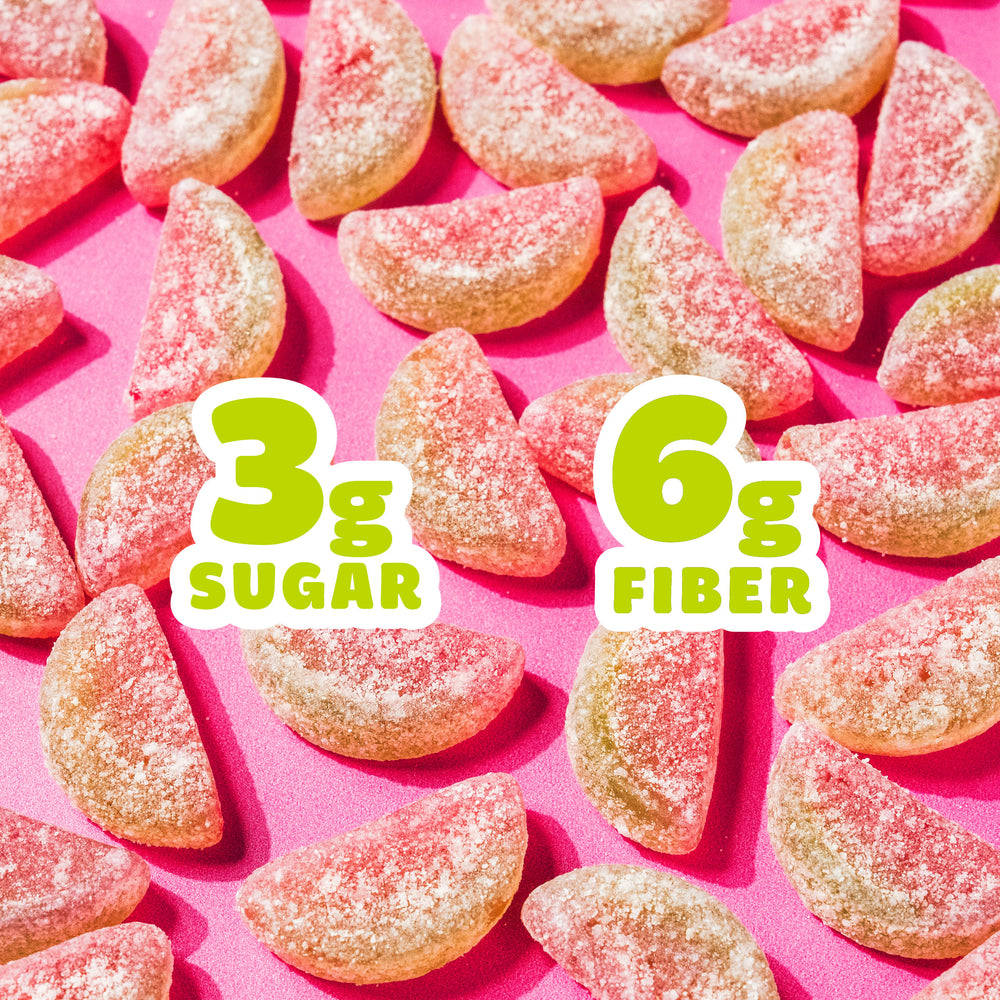 Pile of sour watermelon gummy slices on a pink background. Text overlays read "3g SUGAR" and "6g FIBER".
